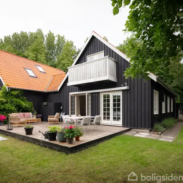 Sortmalet hus med hvid balkon og orange tag, omgivet af grøn have. Der står havemøbler og blomsterkrukker på en terrasse foran huset, der er dækket af græs og træer.