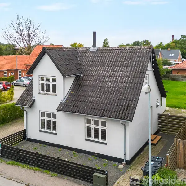 Et hvidt hus med sort tag står stille ved en vej. Det er omgivet af en lav hæk og flisebelagt indkørsel. Nabohuse i rød mursten ses i baggrunden.