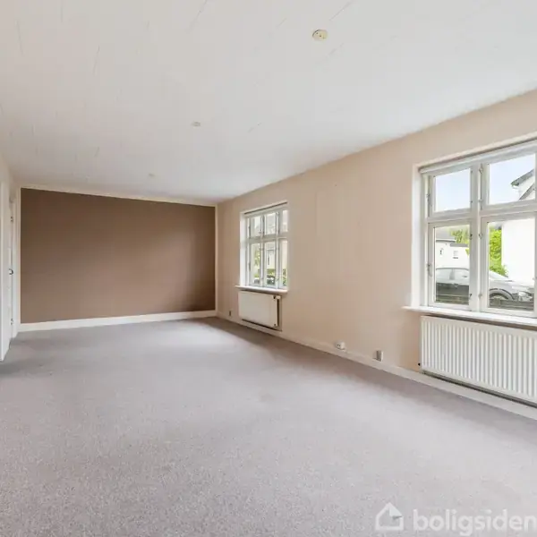 Stuen har beige vægge og gråt gulvtæppe. To store vinduer slipper lys ind, mens en radiator ses under det ene vindue. Værelset er tomt uden møbler.