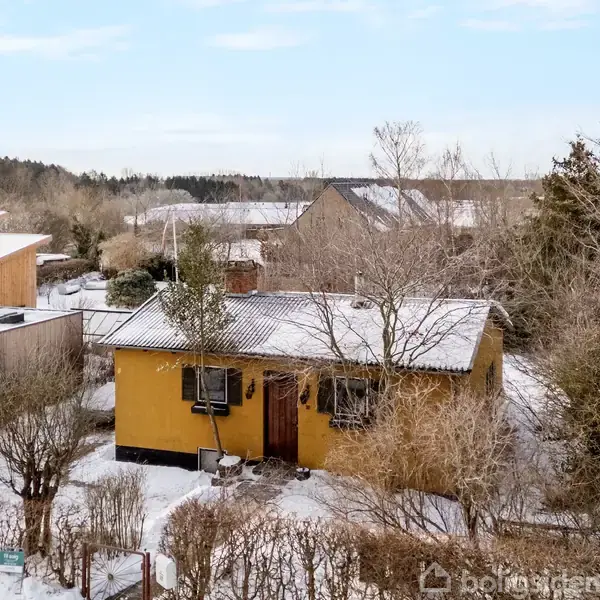 Et lille, gult hus står stille omgivet af sneklædte buske og træer i et roligt nabolag, hvor flere huse ses i baggrunden under en blå himmel.