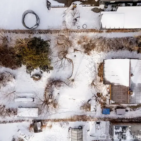 Træer dækket af sne står stille omkring en snedækket have, hvor en bygning med mørkt tag ses. Omgivet af hegn, med stier der skærer gennem sneen.
