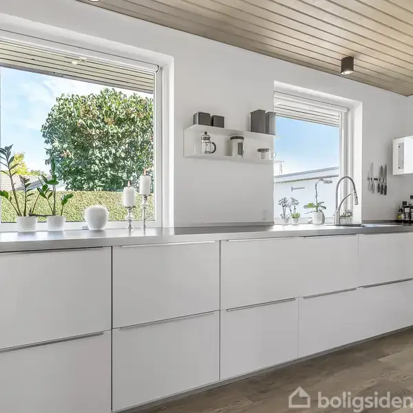 Hvidt køkkenbord i minimalistisk stil med planter og porcelæn. Store vinduer giver udsigt til en grøn have. Moderne indretning med trægulv og skabsplads.