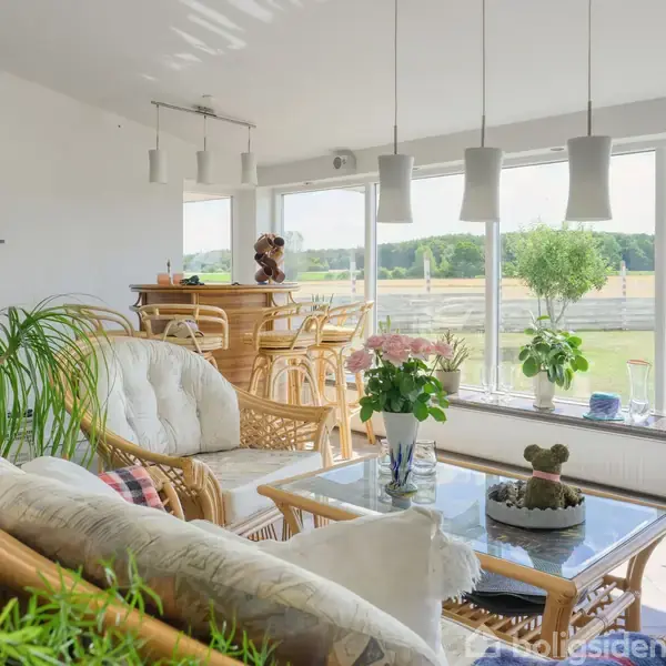 En lys stue med hvide vægge fyldt med planter og kurvemøbler. Et glasbord med en vase af lyserøde blomster står foran en stor vinduesvæg, der viser en grøn have.