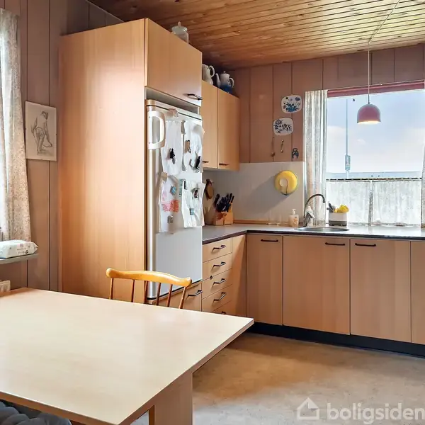 Et køkken med træskabe og lysebrunt interiør. En spisebord står foran et vindue med gardiner. Køleskabet er dekoreret med magneter. Rummet er oplyst af loftslamper.