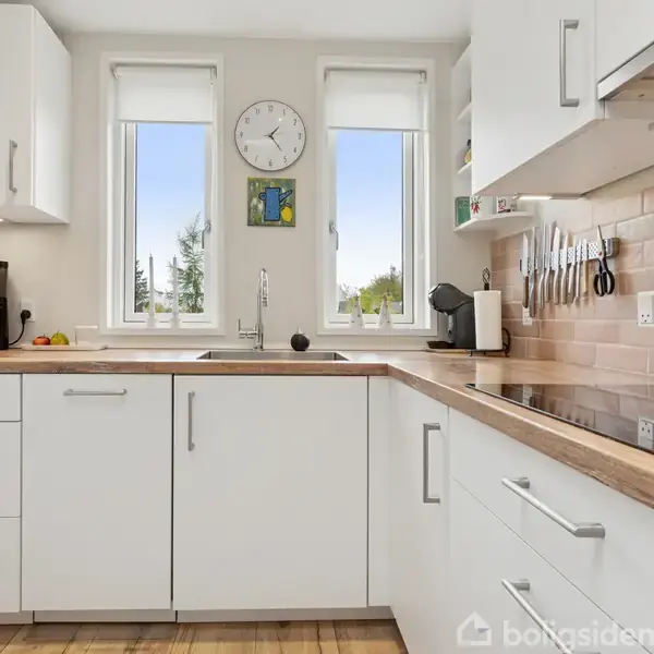 Køkken med hvide skabe, kaffemaskine og vask; knivholder på væggen. To vinduer og et ur viser tid. Trægulv og neutral flisebaggrund skaber en moderne stemning.