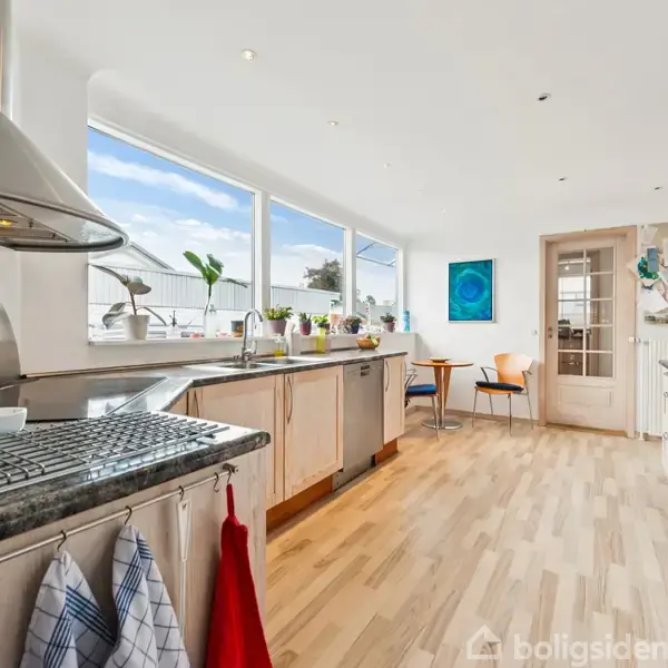 Et moderne køkken med komfur og udtræksemhætte, omgivet af lyse træskabe. Store vinduer lukker dagslys ind og giver udsigt til en terrasse. Et lille bord med stole ses i baggrunden.