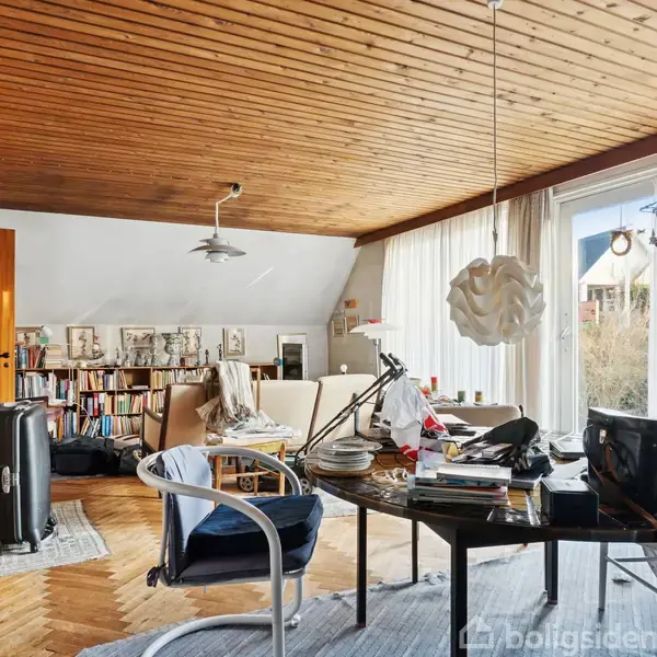 Et rundt bord står i stuen, dækket med bøger og elektronik. Rummet har trægulv og træloft, indrammet af hvide gardiner med naturligt lysindfald. Bogreoler fylder bagvæggen.