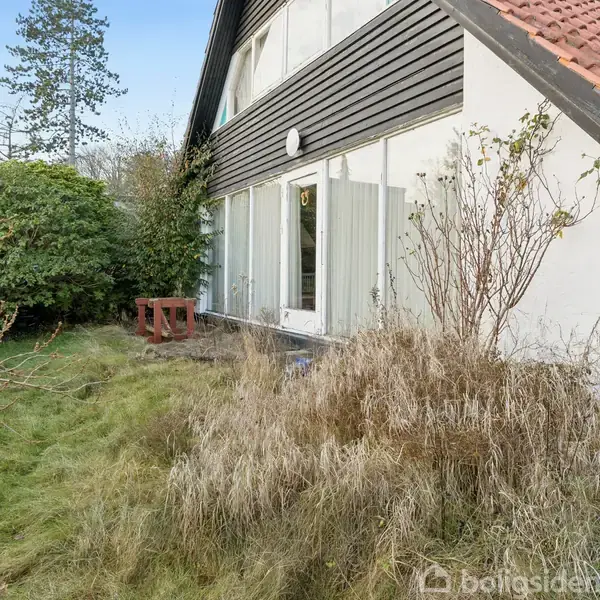 Hus med rød tagsten, store vinduer og havedør. Græs og buske omgiver huset. En bordeaux havebænk står ved siden af, og træer ses i baggrunden under blå himmel.