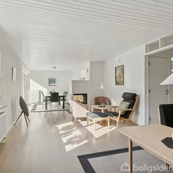 Stue med lyse træmøbler og grønne planter på bord og vindueskarm. Spisebord og stole foran pejs, lysindfald fra store vinduer. Hvide vægge og lofter med ophængt lampe.