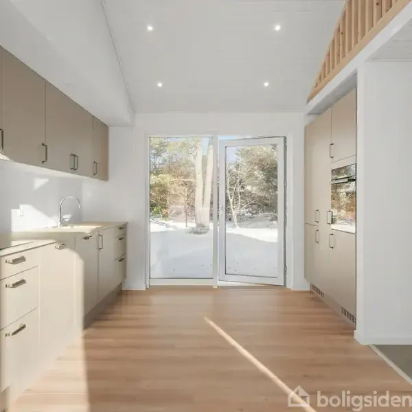 Et lyst køkken med beige skabe og skuffer, der står åbne mod en stor glasdør. Gennem døren ses en snedækket have med træer i baggrunden.