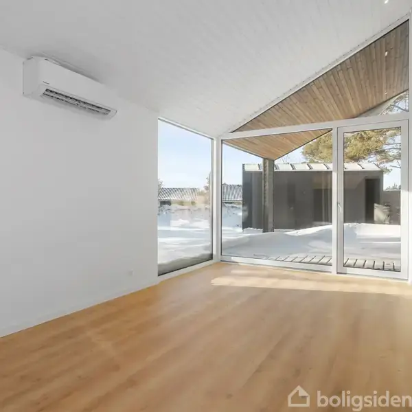 Et lyst rum med et trægulv, en brændeovn til højre og store vinduer, der lader naturligt lys komme ind. Udenfor ses sne og en moderne terrasse.