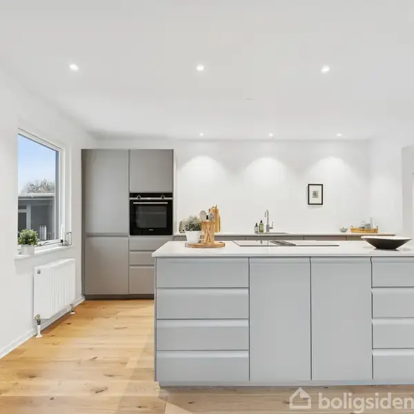 Køkkenø med grå skuffefronter i et moderne køkken, omgivet af hvide vægge og trægulv. En indbygget ovn og vindue med udsigt er i baggrunden.