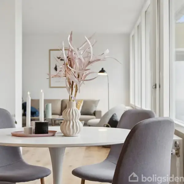 Rundt bord med vase og stearinlys står i en lys spisestue med grå stole. I baggrunden er der en sofa, gulvlampe og vægkunst ved store vinduer.