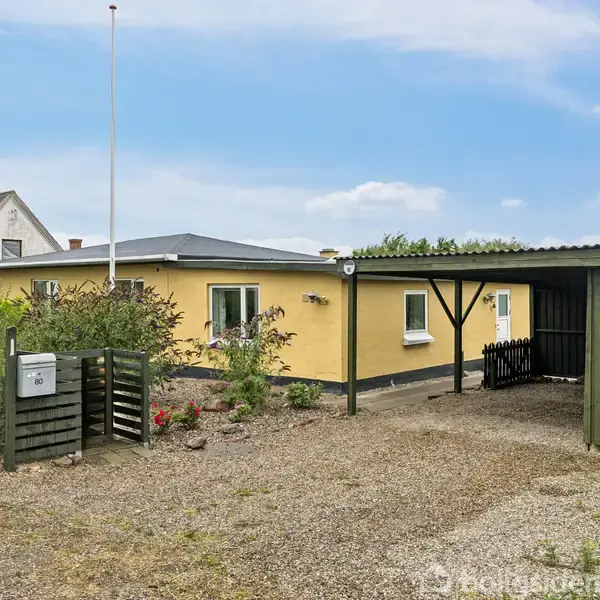 Et gult, enkelt hus står stille på en grusbelagt gårdsplads, omgivet af grønne buske og en carport til højre. En postkasse er placeret nær en lav havelåge.
