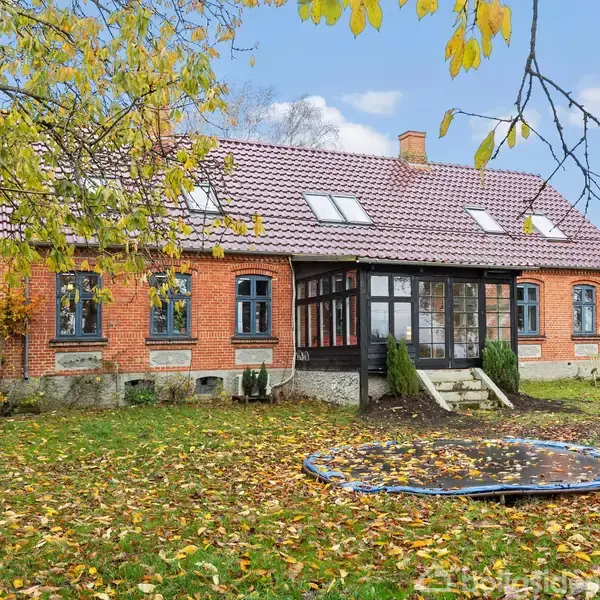 Et rødt murstenshus med skråtag står stille i en have. En trampolin er halvt dækket af faldne blade foran huset, omgivet af træer med gule blade.