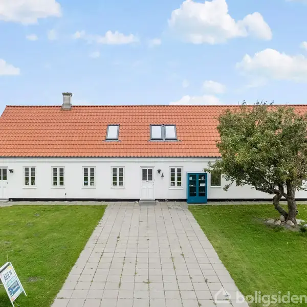Et hvidt hus med rødt tegltag står på en flisebelagt sti omgivet af en græsplæne og et træ. Skyfri blå himmel med enkelte skyer i baggrunden.