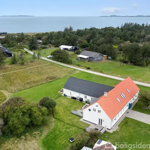 Hus med rødt tag blandt grønne marker ved kysten. Omkringliggende bygninger og stier fører mod en skovbevokset baggrund, og en rolig havudsigt danner horisonten.