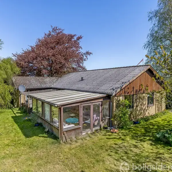En enkel etplanshus med en udestue står på en grøn græsplæne omkranset af træer og buske under en klar blå himmel.