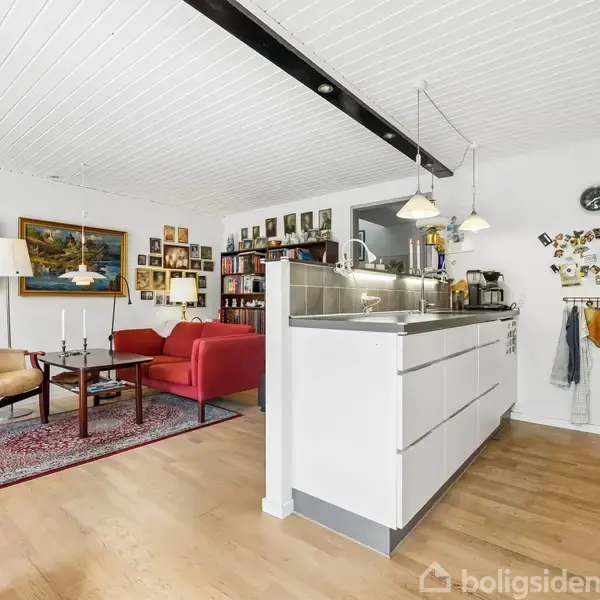 Køkken-alrum med hvidt interiør og pendellamper. Stueområde med rød sofa, lænestol og billeder på væggen. Hylder med bøger i baggrunden. Lyse trægulve og hvidt loft.