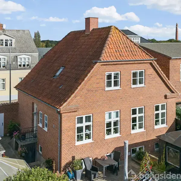 Rødstenshus med tag af tegl, stående på en gårdsplads med havemøbler. Omkringliggende bygninger viser en bysetting med gamle og moderne arkitektoniske stilarter.