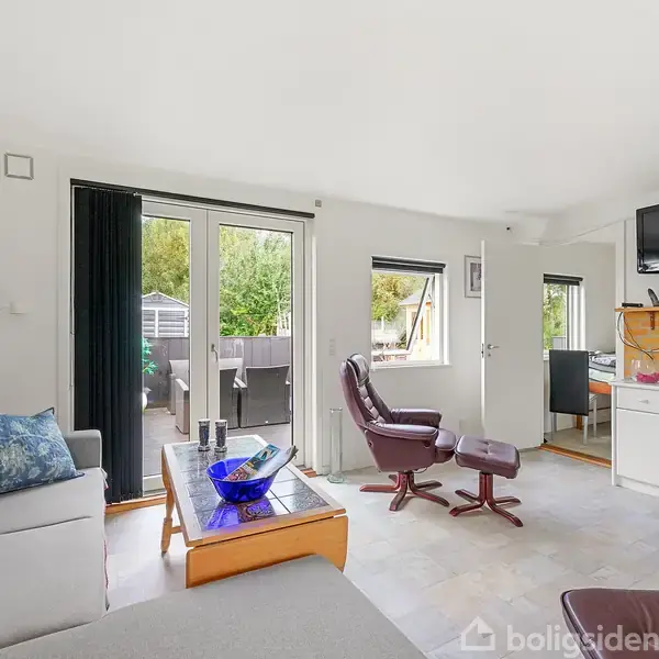 Stue med sofa og lænestol ved et sofabord, store vinduer fører ud til terrasse, lysindfald fra dør til haven. Vægmonteret TV over kaminen, minimalistisk indretning med lyse nuancer.