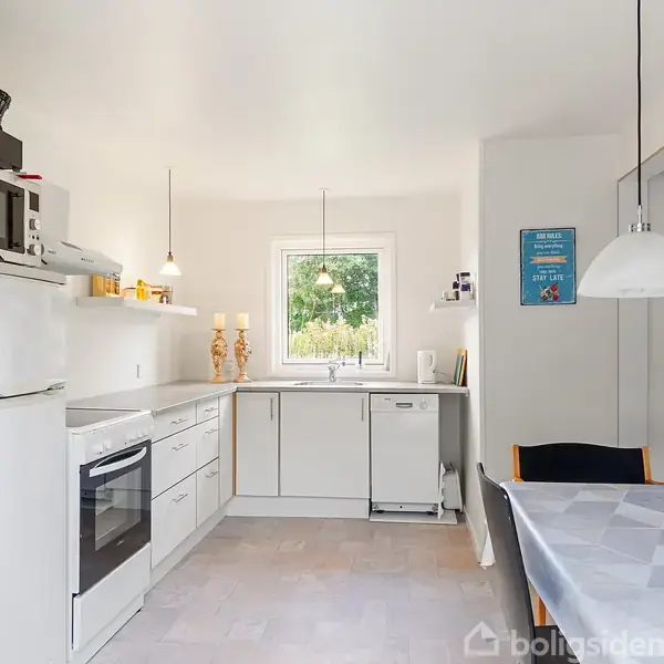 Et lyst køkken med hvidt interiør viser et køleskab, ovn og mikroovn til venstre. Et spisebord med en voksdug og en tændt stearinlys er til højre. Vinduet tilbyder udsigt til grønne omgivelser.