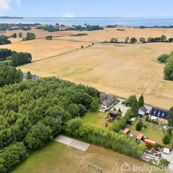 Luftfoto viser en landlig ejendom omgivet af marker og skov. Flere bygninger står i en lille klynge ved en vej, med åbne marker og en fjord i baggrunden under en blå himmel.
