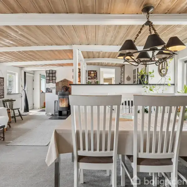 En spisebord med hvide stole står i midten. Ovenover hænger en lysekrone. I baggrunden ses en stue med brændeovn og lænestole. Vinduet til højre giver udsigt til natur.