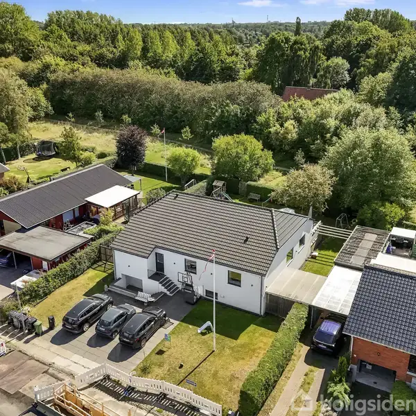 Et hvidt enfamiliehus med gråtag vises fra oven, omgivet af andre huse. Tre biler er parkeret foran. Baggrundens frodige træer giver en grøn og rolig atmosfære.