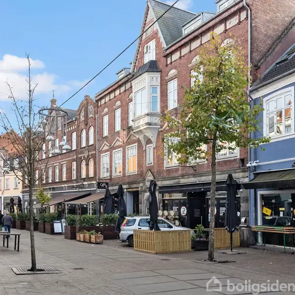 Række af historiske bygninger med butiksgadeniveau, herunder et skilt med teksten "Delici". Biler og træer langs fortovet. Klar himmel og let skydække skaber en lys atmosfære.