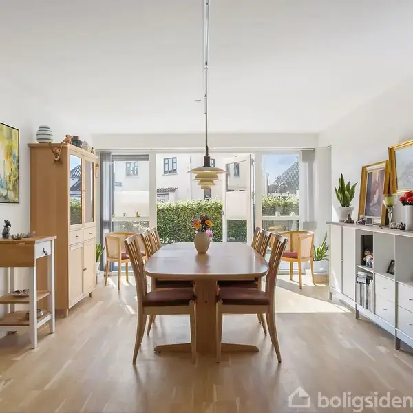 Et spisebord står centralt med blomster i en vase. Rummet er lyst med træmøbler, malerier på væggene og en stor vinduesfront, der viser en have udenfor.