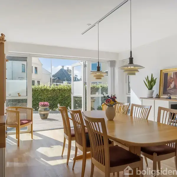 Spisebordet står centralt dækket med en vase, mens stole omkranser det. Rummet har store glasdøre, der åbner ud mod en terrasse og en grøn have.
