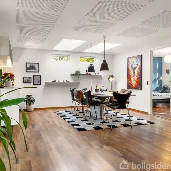 Spisebord med stole står centralt i en moderne spisestue med lyse lofter og trægulv. Rummet er dekoreret med planter, vægkunst og lamper. En åben dør fører til et tilstødende rum.