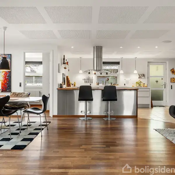 Et moderne køkken integrerer en ø med barstole, omgivet af spiseplads med sort-hvide stole og farverig kunst. Rummets trægulv harmonerer med den luftige, lyse atmosfære.