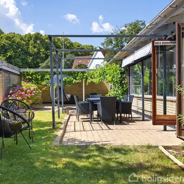 Terrasse med havemøbler står på fliser, omgivet af græs og grønne planter. Åben dør fra huset fører ud til området, der er solbeskinnet med blå himmel og få skyer.