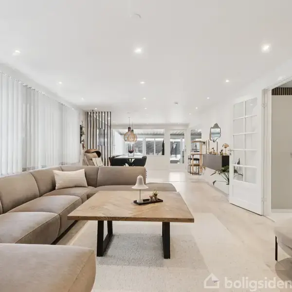 En stor, beige sectional sofa står i en lys stue med et trægulv. Et stort træbord står foran sofaen. Rummet er lyst og moderne med hvide vægge og mange vinduer.