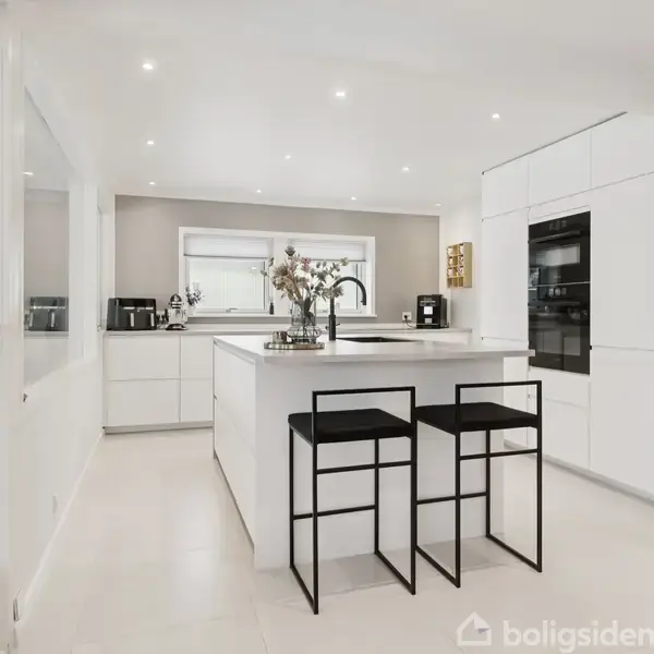 Et moderne køkken med en central ø med to sorte barstole. Køkkenet er lyst med hvidt interiør, indbygningsovn, og blomster på bordet. Store vinduer giver naturligt lys.