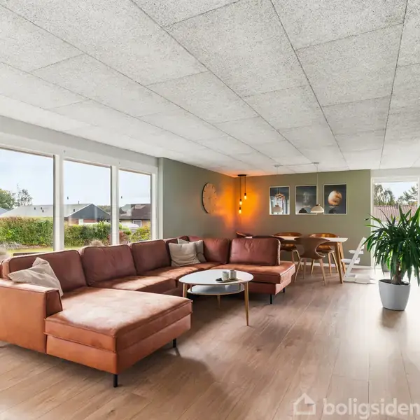 En rummelig stue med en stor, orange hjørnesofa, der står på et trægulv. Et rundt sofabord står foran sofaen. Store vinduer giver udsigt til en have.