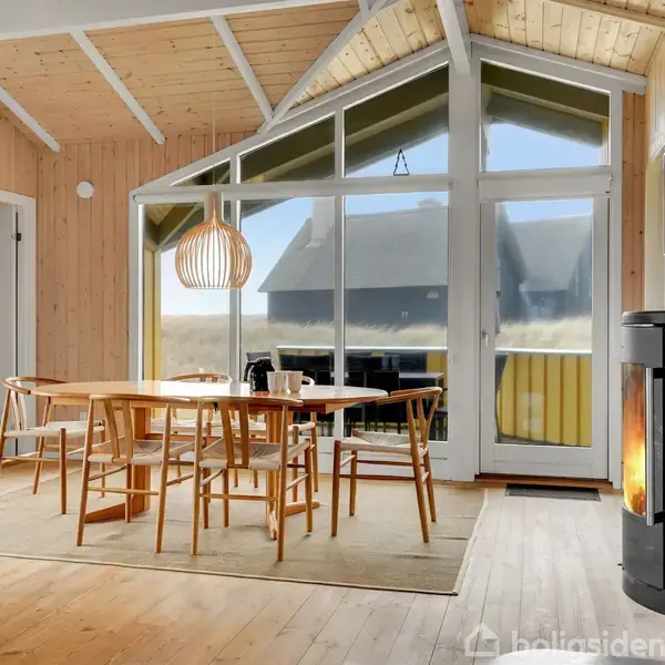 Et spisebord med stole står i en stue med højt til loftet og træpaneler. En moderne pejs brænder, mens store vinduer viser udsigt til natur og en terrasse.