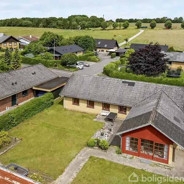 Hus set fra oven med rødt gavlvindue, omgivet af grøn have og flisebelagt terrasse. Nabolaget har flere huse, marker og træer i baggrunden.