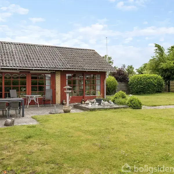 Hus med rødt murværk og store vinduer, har terrasse med bord og stole, omgivet af en grøn have. Et lille springvand står ved siden af terrassen, omgivet af buske og hæk.