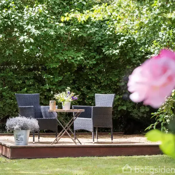 To rattan-stole står ved et rundt bord, dekoreret med blomster, på en lille terrasse. Forgrunden viser en rose, mens omgivelserne er tæt beplantet med grønne buske.