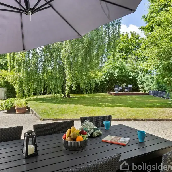 Havebord med kurvestole og skål med frugt står under en stor parasol. I baggrunden ses en grøn have med træer og en terrasse med siddepladser.