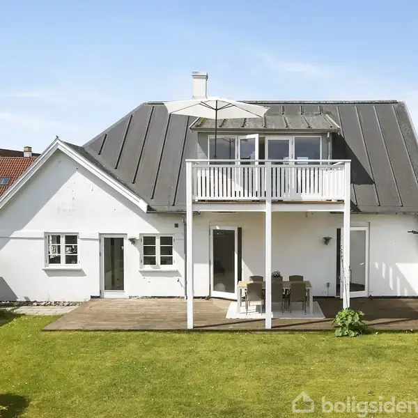Sofagruppe med marmorbord og blomster på tæppe; bagved ses spisebord med stole og hængelampe. Store vinduer lukker lys ind i det lyse, moderne rum med trægulv og kunstværker.