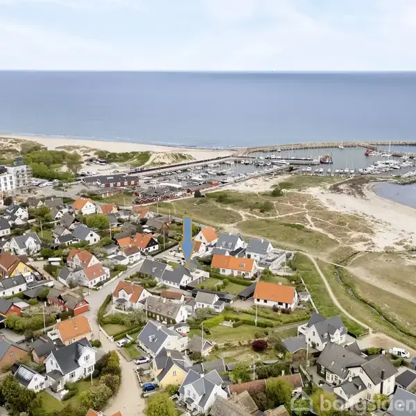Hus med gråt tag og hvide vinduer står stille i en kystnær landsby. Haven er velplejet, og der er udsigt til andre huse og havet i baggrunden.