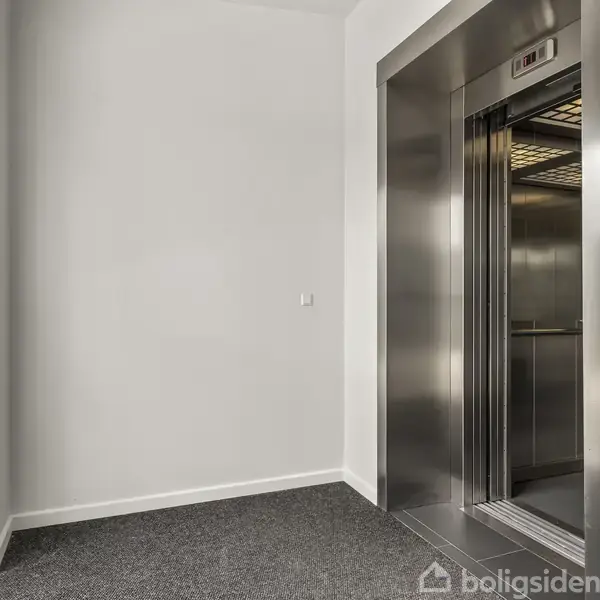 Elevator står åben i en lys gang med gråt gulvtæppe og hvidmalede vægge. Til venstre er der et vindue, der lukker naturligt lys ind.