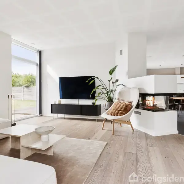 Stue med hvid sofa, fladskærms-TV på væggen, moderne pejs, og grøn plante. Store vinduer slipper naturligt lys ind. Spisebord og stole ses i baggrunden ved åbent køkken.