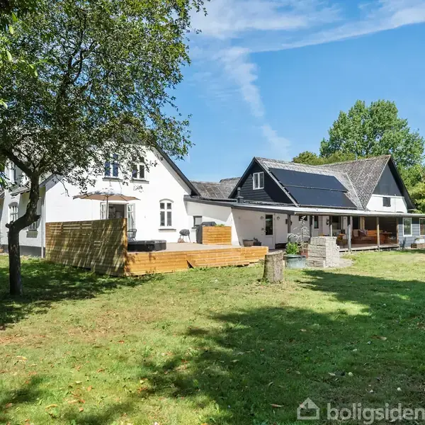 Hus med solpaneler på taget står stille i grøn have med flere træer omkring. Blå himmel og græsplæne dominerer omgivelserne.