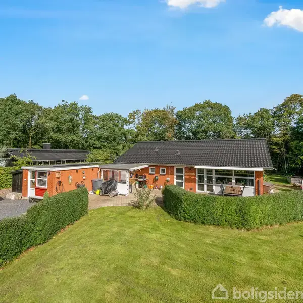 Et rødt hus med sort tag står i en grøn have med hæk omkring terrassen. I baggrunden ses tæt bevokset skov. Himmelen er blå med få skyer.
