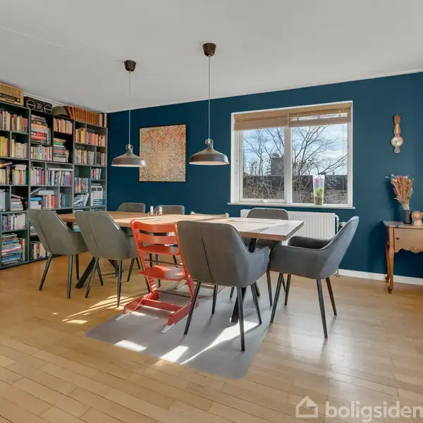 Stue med spisebord og seks stole, hvoraf én er rød. Hylder fyldt med bøger står langs væggen. Blåtmalet væg med kunst, to loftslamper og vindue med træer udenfor.